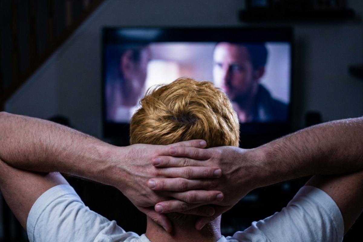 Osoba sedi ispred televizora i gleda film ili seriju u zamračenoj prostoriji, opuštena atmosfera kućnog gledanja sadržaja.